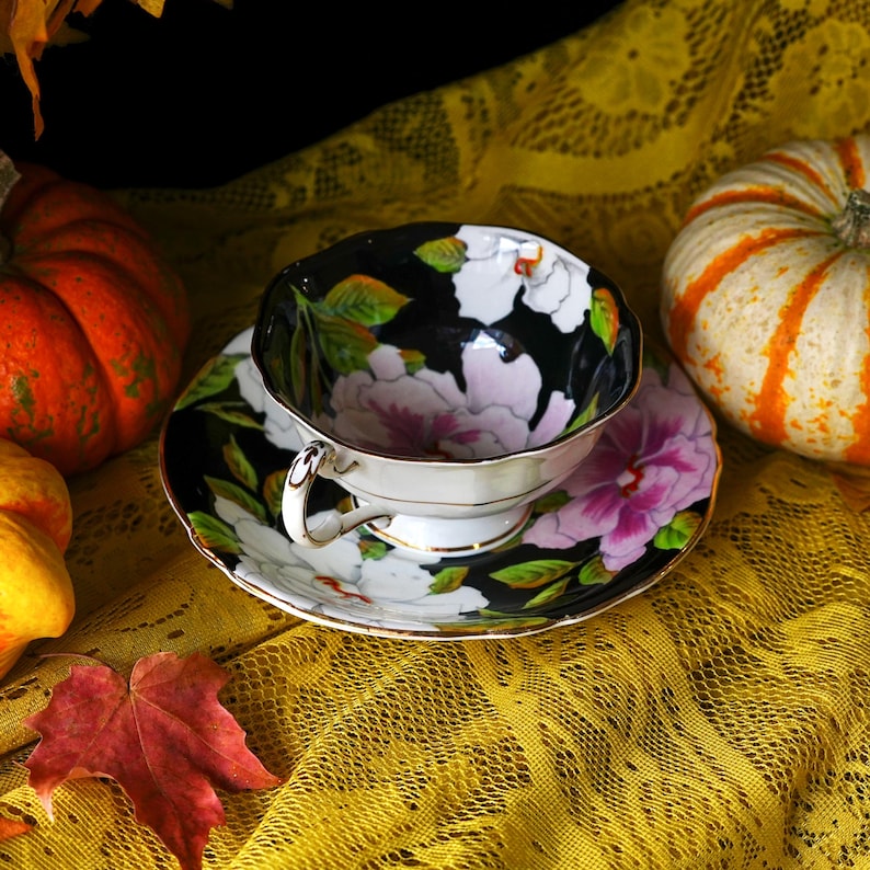 May include: A black teacup and saucer set with a floral design featuring white and pink flowers and green leaves. The teacup and saucer are on a yellow lace tablecloth, with pumpkins in the background.