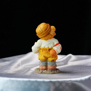 Op de afbeelding: Een klein beeldje van een beer met een gele hoed, wit shirt, gele broek en grijze en roze laarzen. De beer staat op een kleine, getextureerde basis. De beer staat met zijn rug naar de kijker.