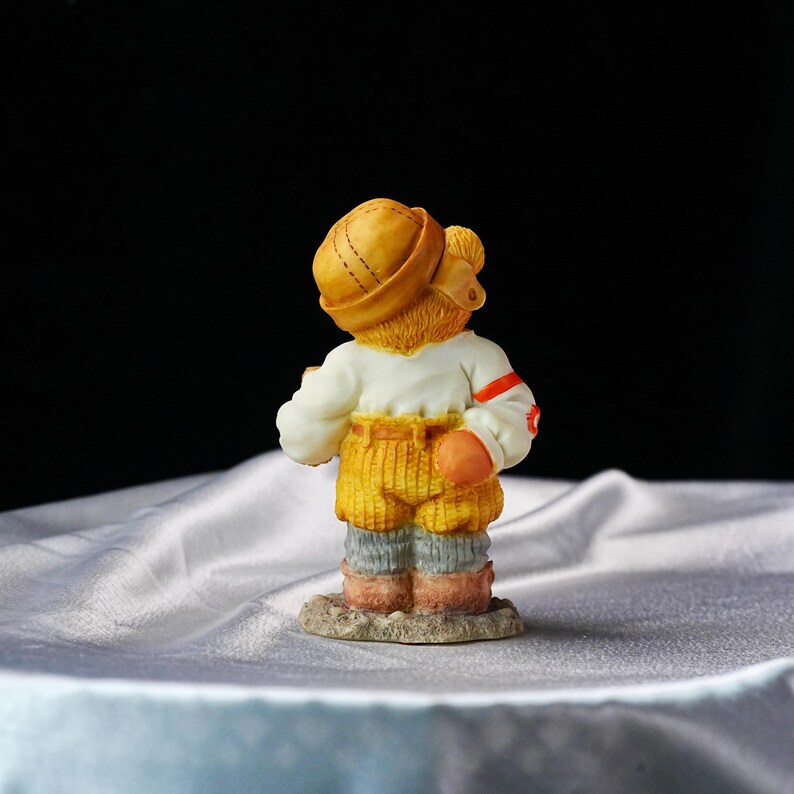 Op de afbeelding: Een klein beeldje van een beer met een gele hoed, wit shirt, gele broek en grijze en roze laarzen. De beer staat op een kleine, getextureerde basis. De beer staat met zijn rug naar de kijker.