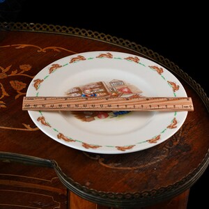 May include: A white ceramic plate featuring a central illustration of a rabbit in a shop labeled 'Mr. Piggly's Stores' and a border of running rabbits. A wooden ruler placed across the plate shows a diameter of approximately 8 inches.