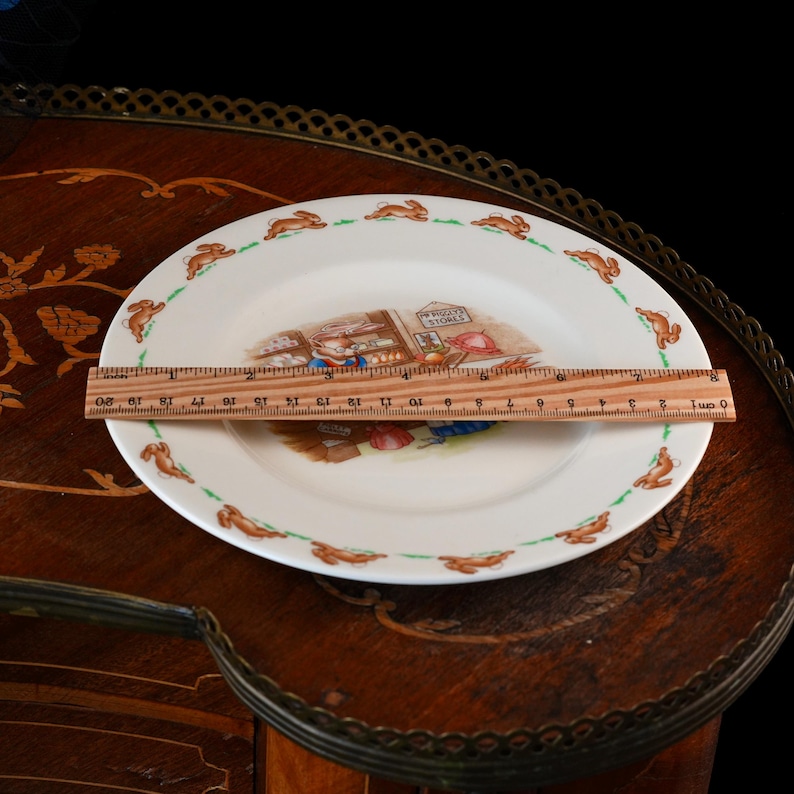 May include: A white ceramic plate featuring a central illustration of a rabbit in a shop labeled 'Mr. Piggly's Stores' and a border of running rabbits. A wooden ruler placed across the plate shows a diameter of approximately 8 inches.