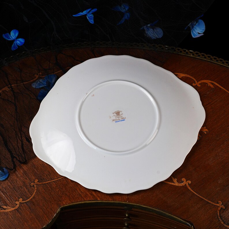 Puede incluir: Un plato de porcelana blanca con borde festoneado, visto desde la parte inferior. El centro presenta una peque&ntilde;a marca de fabricante con el texto Royal Albert, Bone China, England y el nombre del patr&oacute;n Albany Blue, impreso en tinta negra y azul.