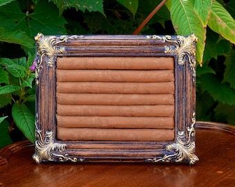 May include: A rectangular, brown jewelry display with a gold and dark brown ornate frame. The display has multiple horizontal slots for rings or other small jewelry items. The background is a blurred view of green foliage.
