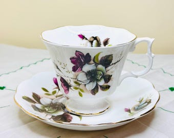 May include: A white teacup and saucer with a floral design. The flowers are white, purple, and green. The cup and saucer have a gold trim.