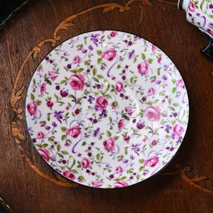 May include: A white porcelain saucer featuring a floral design of pink roses, purple details, and green foliage. The saucer has a black rim and is placed on a dark wooden surface. The pattern is a classic, vintage style.