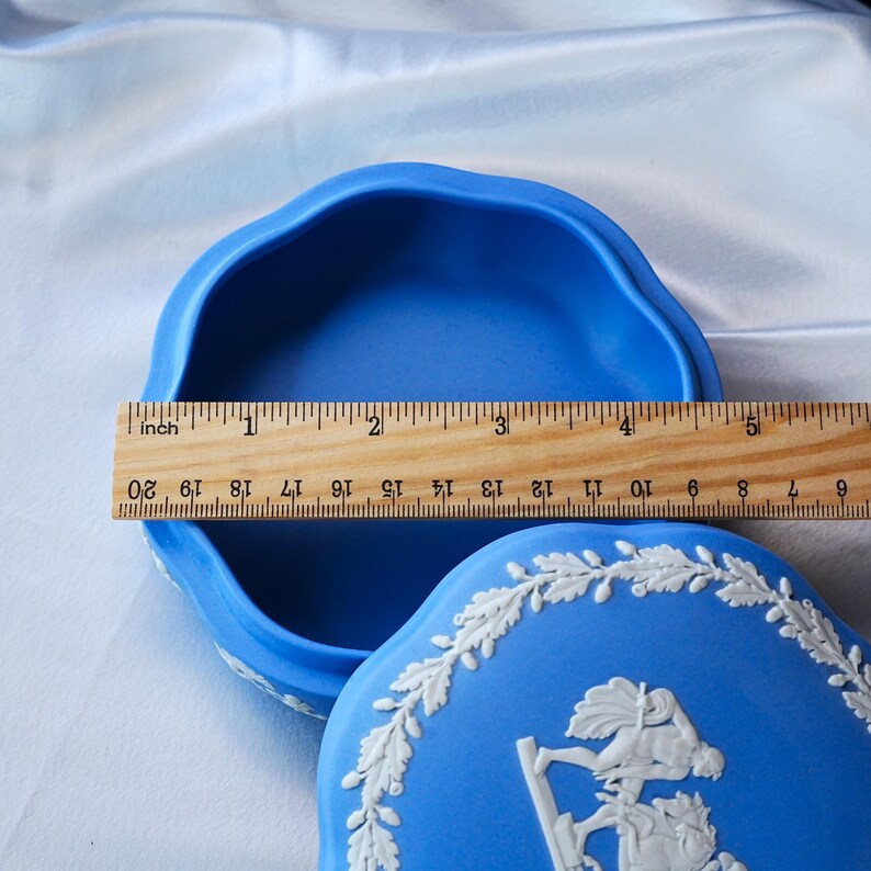 May include: A blue round lidded box with white relief detailing featuring a figure and a wreath pattern. A wooden ruler is placed across the open container, showing a diameter of approximately 5 inches for the interior opening of the vessel.