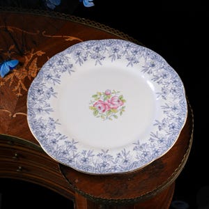 Puede incluir: Un plato redondo de cer&aacute;mica blanca con borde festoneado, decorado con un patr&oacute;n floral azul y un ramillete central de rosas rosas con hojas verdes. El borde del plato est&aacute; rematado con un fino ribete dorado que a&ntilde;ade un toque decorativo.