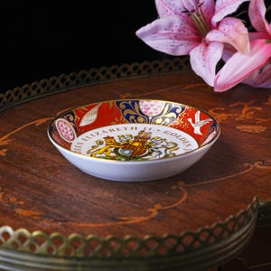 May include: A round porcelain dish featuring a commemorative design for the Queen Elizabeth II Golden Jubilee. The interior displays a royal coat of arms with a lion and unicorn, surrounded by red, blue, and gold patterns with white bird motifs.