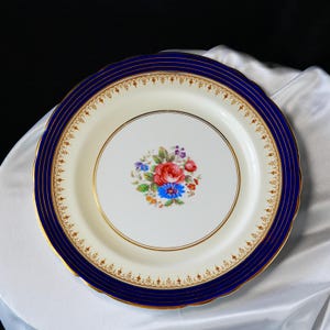 Pode incluir: Um prato decorativo de porcelana com centro branco e borda azul marinho. A borda apresenta detalhes dourados e um desenho floral. O centro do prato mostra um buqu&ecirc; de rosas vermelhas, cent&aacute;ureas azuis e outras flores coloridas.