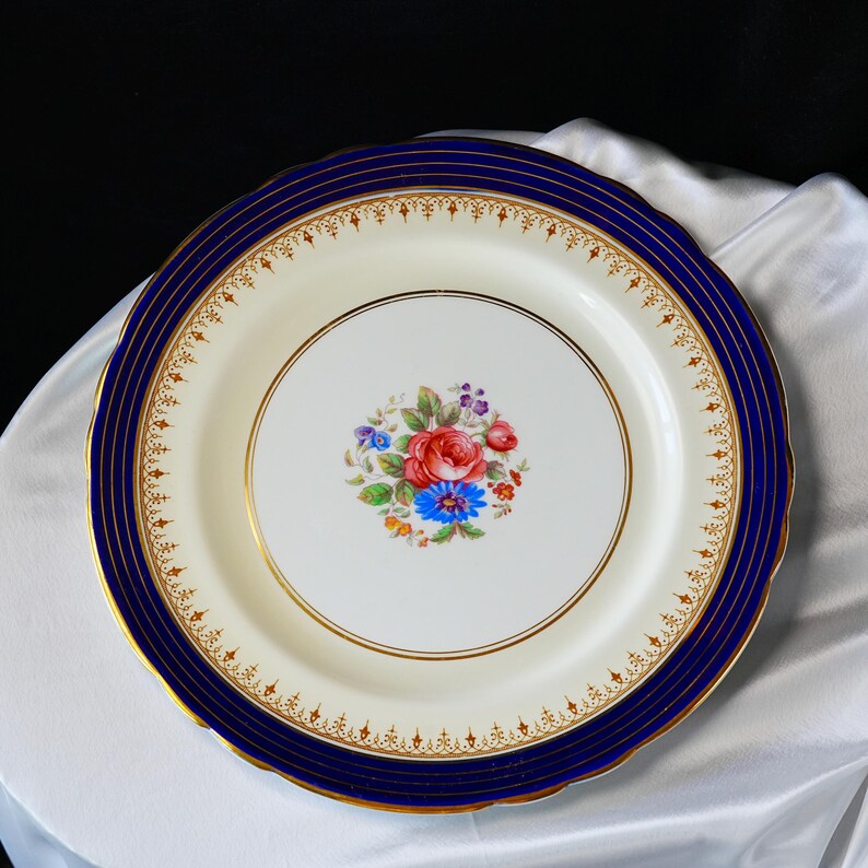 Pode incluir: Um prato decorativo de porcelana com centro branco e borda azul marinho. A borda apresenta detalhes dourados e um desenho floral. O centro do prato mostra um buqu&ecirc; de rosas vermelhas, cent&aacute;ureas azuis e outras flores coloridas.
