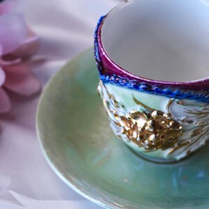 May include: A detailed close-up of a vintage teacup and saucer set. The cup features a white interior, a purple and blue rim, and gold floral detailing. The saucer and the cup's exterior are a soft green.