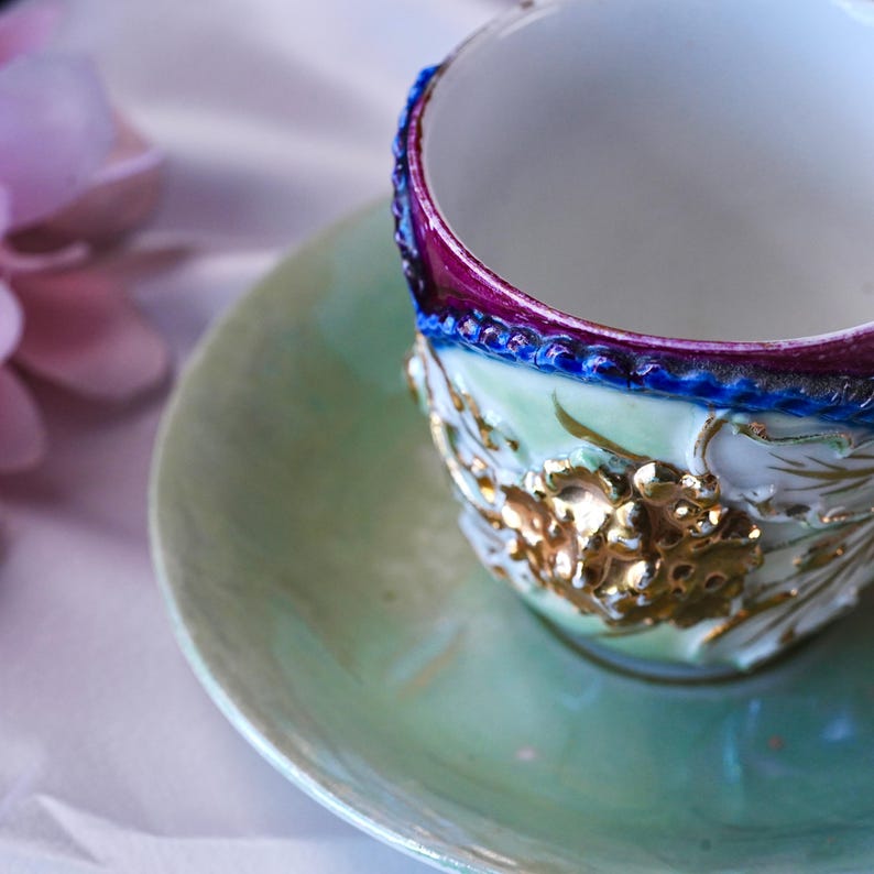 May include: A detailed close-up of a vintage teacup and saucer set. The cup features a white interior, a purple and blue rim, and gold floral detailing. The saucer and the cup's exterior are a soft green.