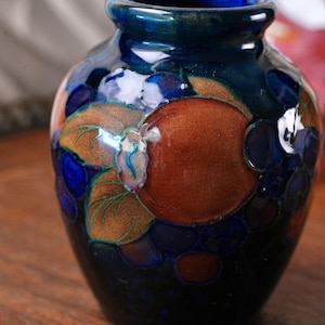 May include: A small, rounded ceramic vase with a glossy, deep blue glaze. The surface features raised, embossed relief patterns of round fruit and leaves in shades of copper, bronze, and dark blue, creating a textured, multi-dimensional appearance.