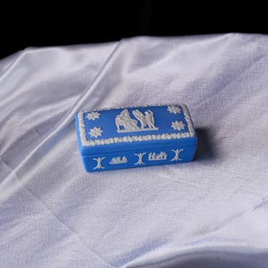 May include: A blue rectangular trinket box with white relief detailing. The box features floral and classical figures around the edges and on the lid. The box is resting on a white, silky surface.