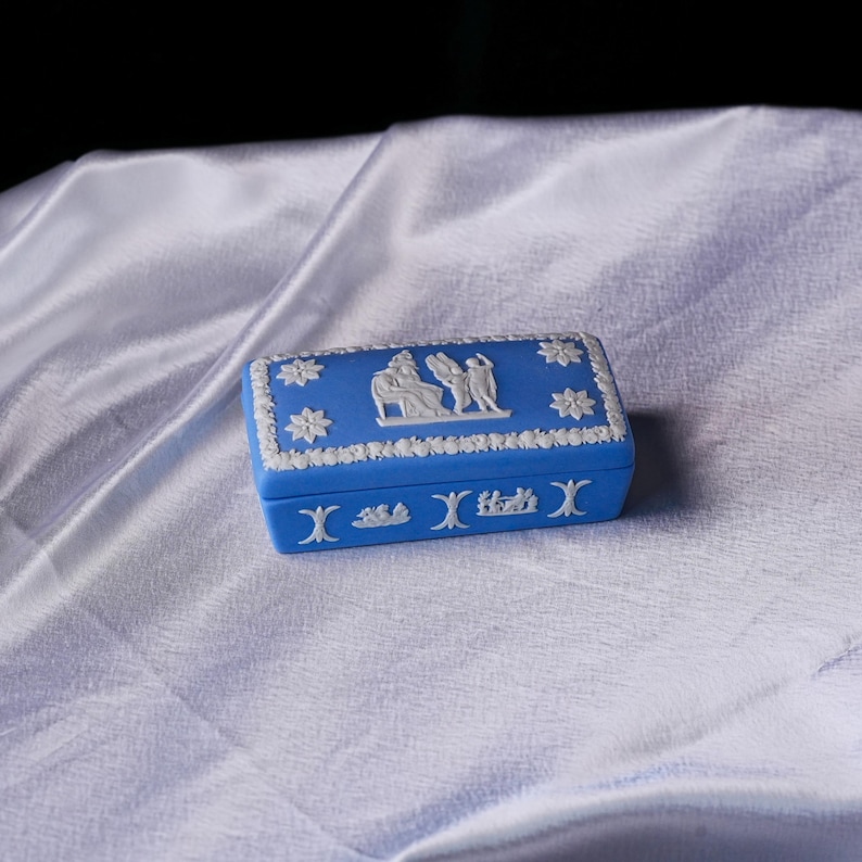 May include: A blue rectangular trinket box with white relief detailing. The box features floral and classical figures around the edges and on the lid. The box is resting on a white, silky surface.