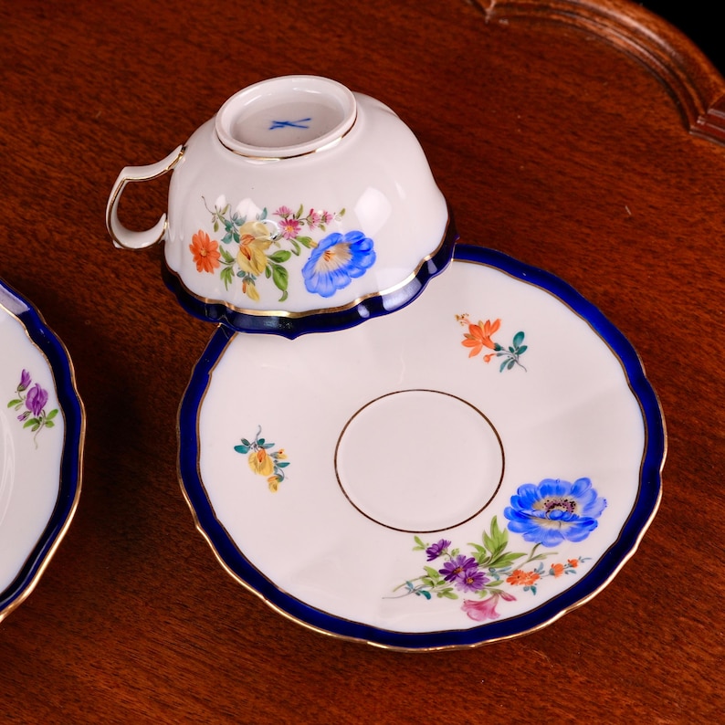 Puede incluir: Juego de taza y platillo de porcelana blanca con borde azul y dise&ntilde;o floral. La taza est&aacute; adornada con flores coloridas, incluyendo azul, naranja y amarillo. El platillo tiene un dise&ntilde;o a juego con un borde dorado.