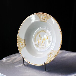 May include: White ceramic bowl with a yellow floral design in the center and on the rim. The rim has a gold checkered pattern. The bowl is on a black metal stand, set against a black background.