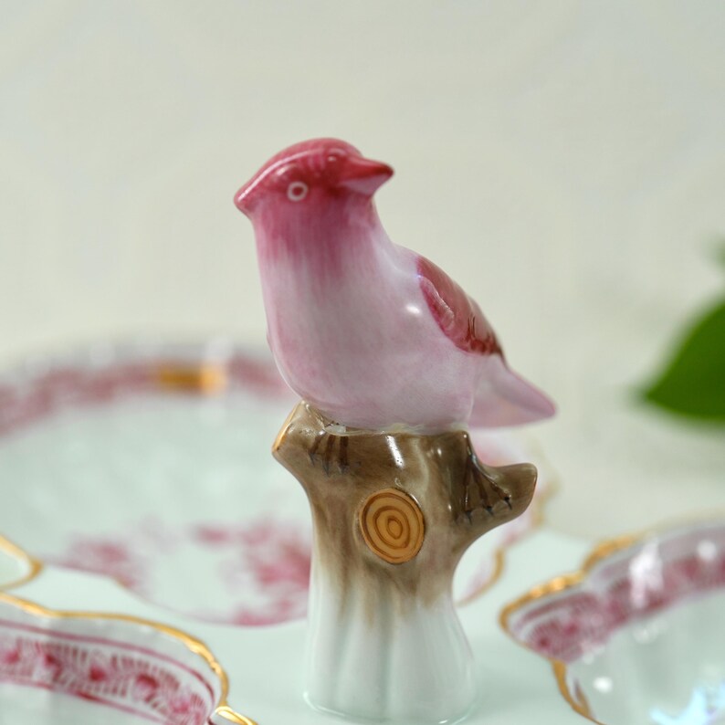 May include: A ceramic bird figurine in shades of pink and red perched on a brown tree stump. The bird has a detailed design and is part of a decorative set. The background includes a glimpse of a matching plate with a floral pattern.