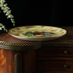 May include: A decorative, vintage-style plate with a floral design. The plate features a gold rim and a painted image of purple grapes, green leaves, and blue flowers. The background is a mix of yellow and brown hues. The plate is set on a wooden surface.