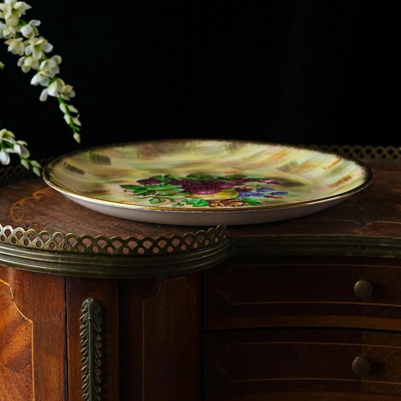 May include: A decorative, vintage-style plate with a floral design. The plate features a gold rim and a painted image of purple grapes, green leaves, and blue flowers. The background is a mix of yellow and brown hues. The plate is set on a wooden surface.