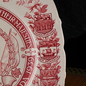 May include: A cream-coloured ceramic plate with a scalloped edge featuring red transferware designs. The text reads "THEIR MAJESTIES" and "CANADA 1939". It displays the coats of arms for Saskatchewan and New Brunswick, surrounded by floral motifs.