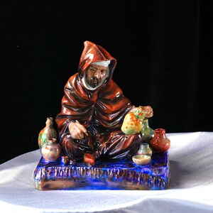 May include: A ceramic figurine of a robed figure with a hood, seated on a blue base. The figure is surrounded by small, colourful vases and a bag. The figurine is painted with shades of red, brown, and blue.