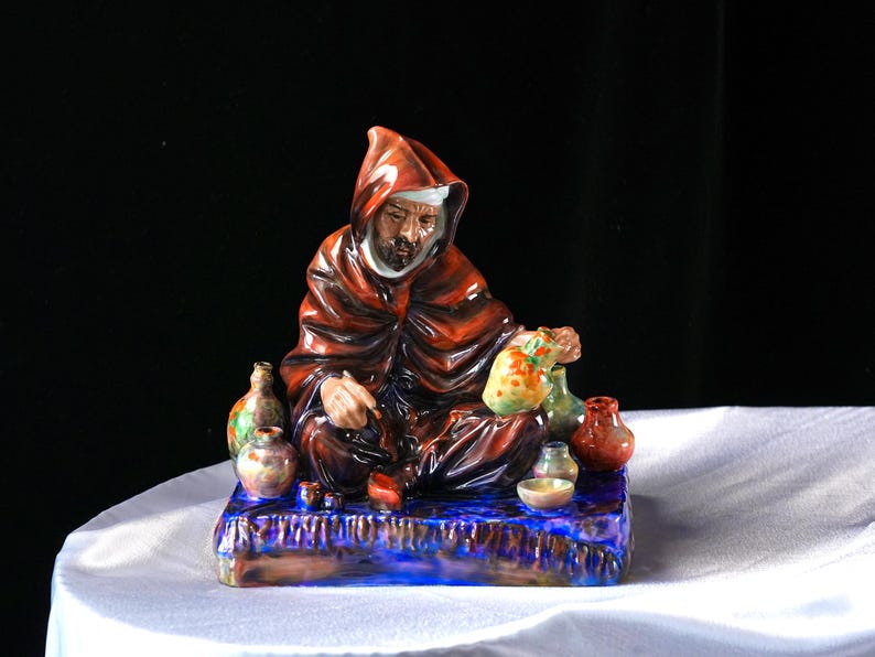 May include: A ceramic figurine of a robed figure with a hood, seated on a blue base. The figure is surrounded by small, colourful vases and a bag. The figurine is painted with shades of red, brown, and blue.
