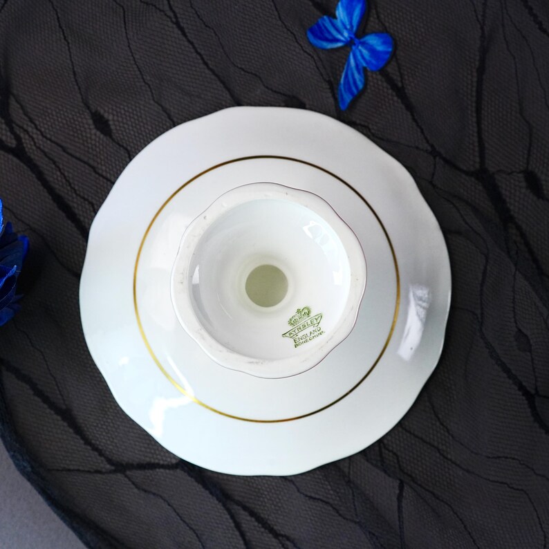 Puede incluir: Un plato de pedestal de cer&aacute;mica blanca con borde festoneado y un fino borde dorado. La parte inferior presenta un sello verde con un icono de corona y el texto "Stanley England Bone China". El plato se ve desde arriba, mostrando su forma circular.
