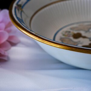 May include: A white ceramic bowl with a gold rim and decorative blue and gold accents. The bowl has a fluted interior and a floral design. The bowl is likely vintage or antique.