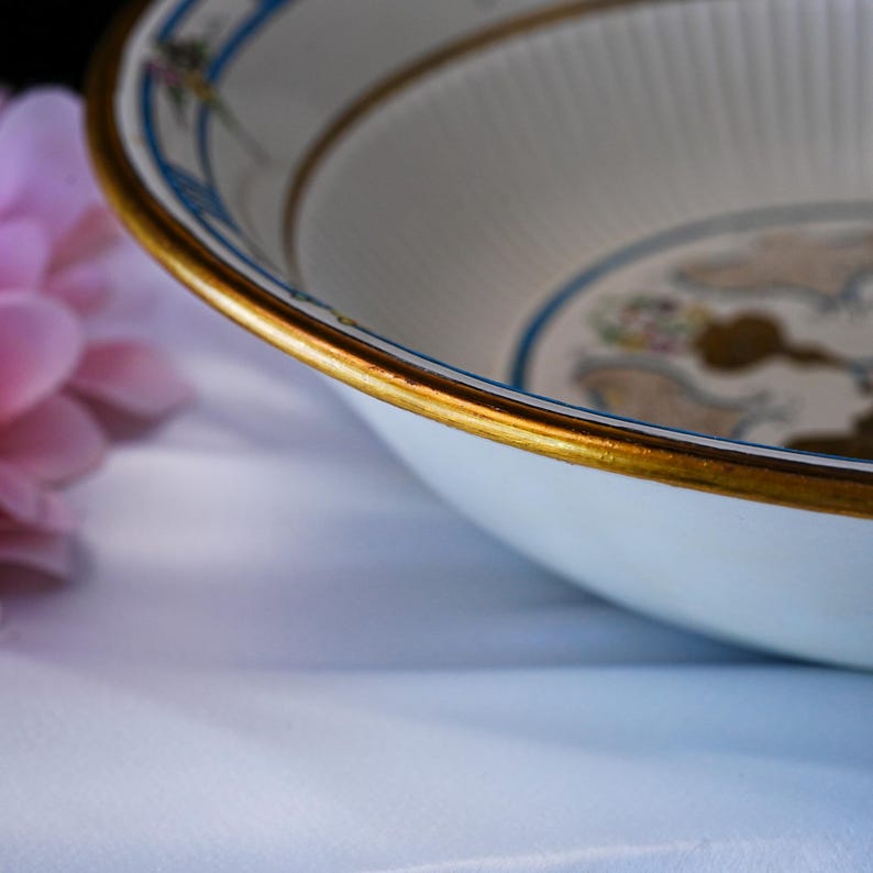 May include: A white ceramic bowl with a gold rim and decorative blue and gold accents. The bowl has a fluted interior and a floral design. The bowl is likely vintage or antique.