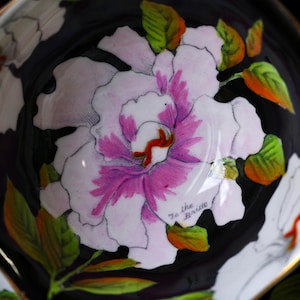 May include: Close-up of a teacup interior with a large, hand-painted floral design. The flower has white petals with pink and purple accents, green leaves, and a red center. The words "To the Bride" are written on the cup.