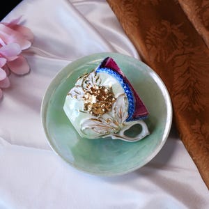 May include: A vintage teacup and saucer set. The cup is pale green with gold floral accents and a gold handle. The saucer is also pale green. The cup is decorated with gold and white floral designs. The rim of the cup is decorated with blue and red trim.