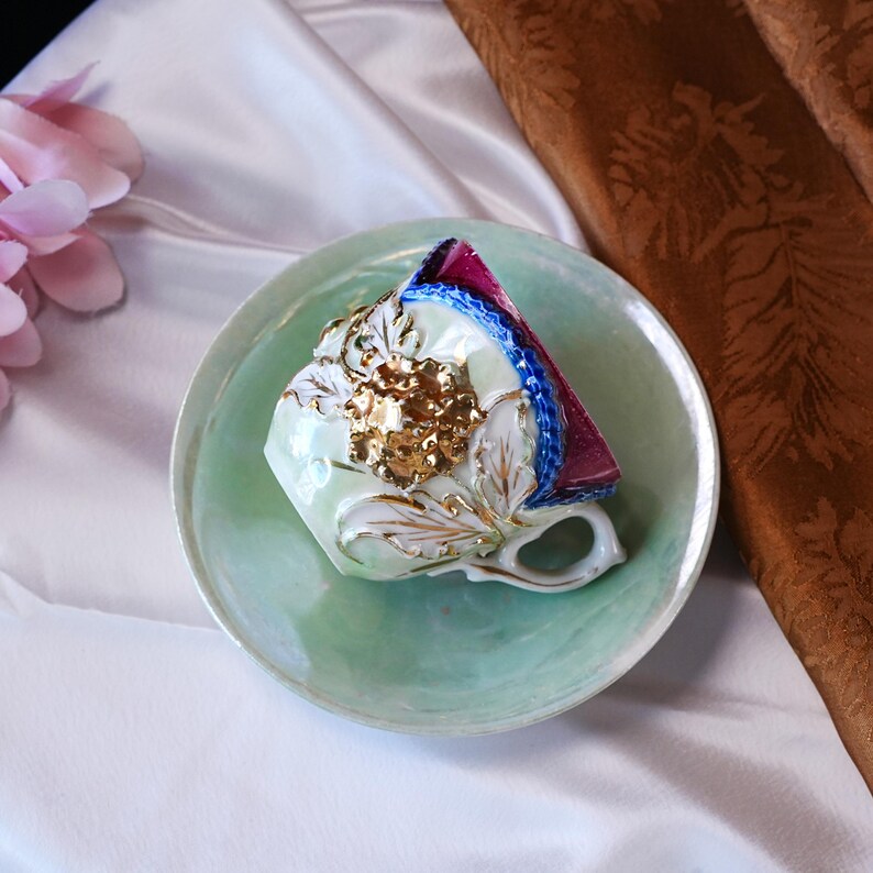 May include: A vintage teacup and saucer set. The cup is pale green with gold floral accents and a gold handle. The saucer is also pale green. The cup is decorated with gold and white floral designs. The rim of the cup is decorated with blue and red trim.
