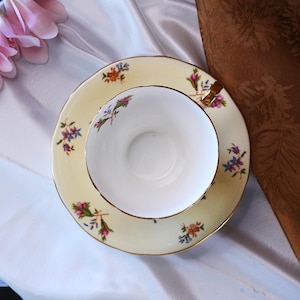 Op de afbeelding: Een vintage theekop en schotel set. De kop en schotel zijn lichtgeel met gouden randen en bloemaccenten in roze, paars en oranje. De binnenkant van de theekop is wit.