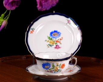 May include: A white porcelain teacup, saucer, and plate set. The set features a blue floral design with gold trim. The plate has a scalloped edge and a dark blue border. The teacup has a handle and a matching floral design.