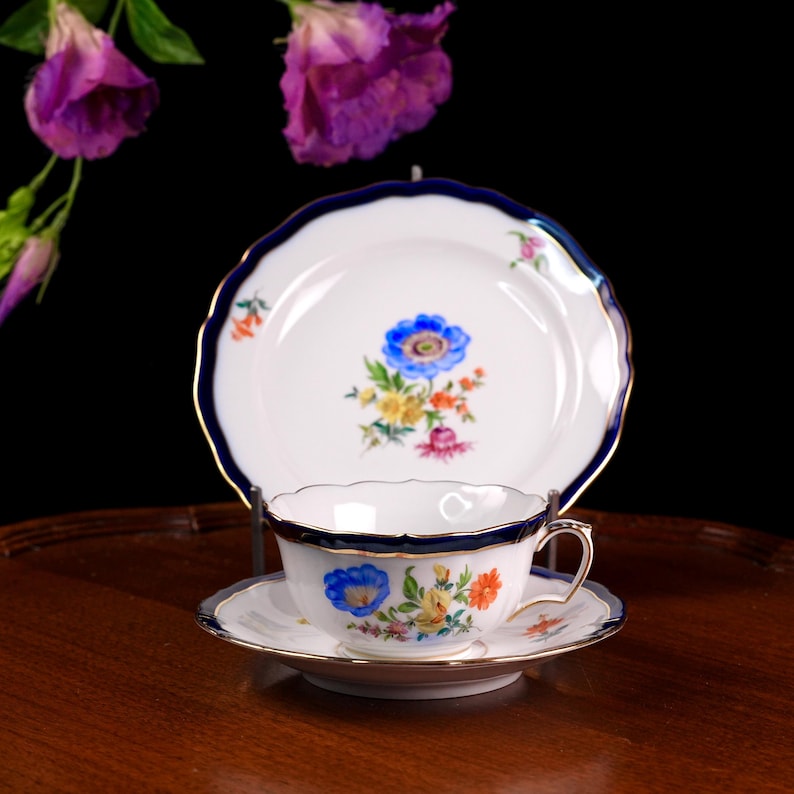 Puede incluir: Juego de t&eacute; de porcelana blanca con taza, platillo y plato. El juego presenta un dise&ntilde;o floral azul con ribetes dorados. El plato tiene un borde festoneado y un borde azul oscuro. La taza tiene un asa y un dise&ntilde;o floral a juego.