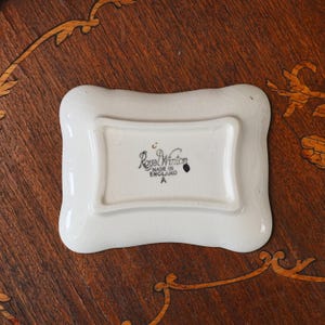 Puede incluir: Un plato rectangular de cer&aacute;mica color blanco hueso con borde festoneado y acabado de esmalte brillante con efecto craquelado. La base tiene un sello negro que dice Royal Winton, MADE IN ENGLAND y la letra A, dentro de un borde elevado.