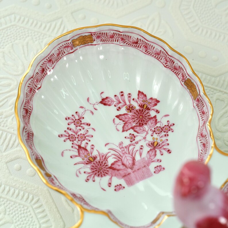 May include: A white porcelain shell-shaped dish with a floral design in shades of pink and gold trim. The dish has a scalloped edge and a detailed floral pattern in the centre. The rim is decorated with a pink and gold border.