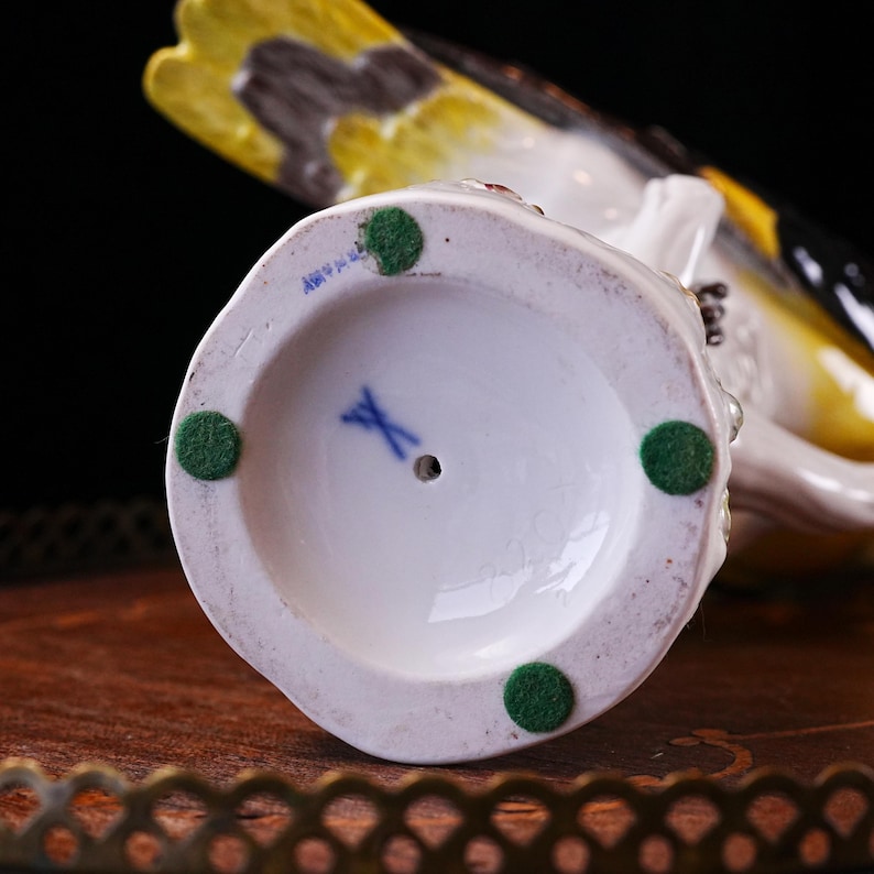 May include: A white porcelain bird figurine with yellow, black, and brown accents. The underside of the figurine is visible, showing a blue logo and green felt pads. The bird is perched on a decorative tray.