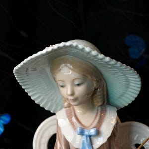 K&ouml;nnte beinhalten: Eine Porzellanfigur einer jungen Person mit einem hellblauen Hut mit breiter Krempe und plissiertem Rand. Die Figur hat hellbraunes Haar und tr&auml;gt ein braunes Kleidungsst&uuml;ck mit wei&szlig;em Kragen und einer kleinen, hellblauen Schleife am Ausschnitt.