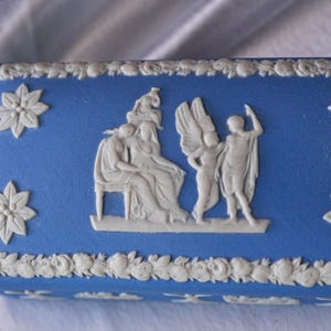 May include: A blue rectangular box with white relief detailing. The box features classical figures and floral patterns. The relief includes a seated woman, winged figures, and decorative floral elements. The box has a matte finish.