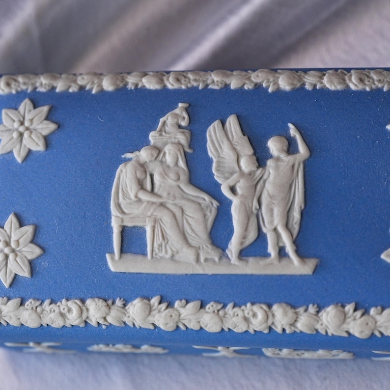 May include: A blue rectangular box with white relief detailing. The box features classical figures and floral patterns. The relief includes a seated woman, winged figures, and decorative floral elements. The box has a matte finish.