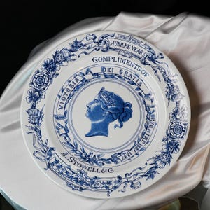 May include: White ceramic plate with blue floral and scrollwork border. The plate features a blue profile of Queen Victoria in a central medallion, with text that reads "Jubilee Year" and "Compliments of Dei Gratia".