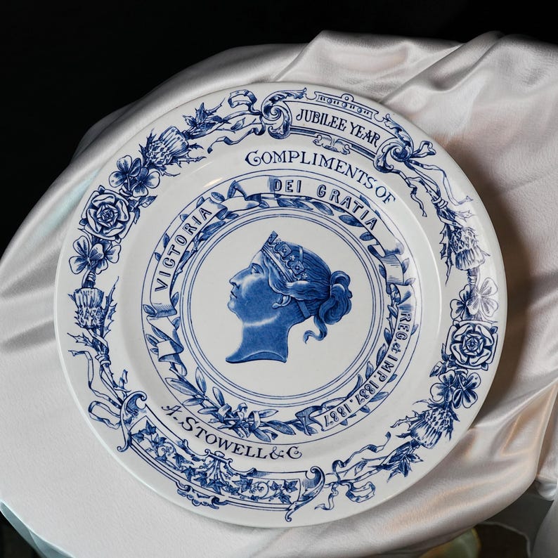 May include: White ceramic plate with blue floral and scrollwork border. The plate features a blue profile of Queen Victoria in a central medallion, with text that reads "Jubilee Year" and "Compliments of Dei Gratia".