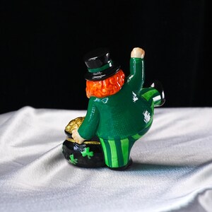 May include: A ceramic leprechaun figurine in a green coat and striped pants, holding a pot of gold. The figurine has a red beard, a black top hat, and a green shamrock on the pot.