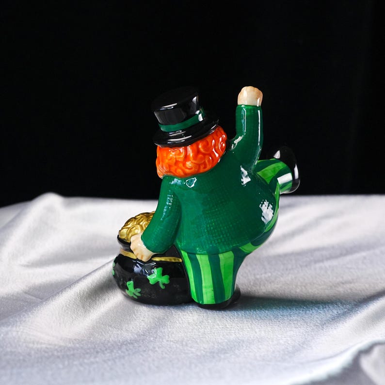 May include: A ceramic leprechaun figurine in a green coat and striped pants, holding a pot of gold. The figurine has a red beard, a black top hat, and a green shamrock on the pot.