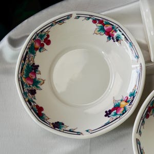Może przedstawiać: Biały ceramiczny spodek z dekoracyjną obw&oacute;dką z kolorowymi owocami i liśćmi. Obrzeże ma czarno-granatowy geometryczny wz&oacute;r. Spodek jest częścią zestawu naczyń.
