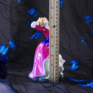 May include: A porcelain figurine of a person in a vibrant magenta and blue gown with a white ruffled hem, holding a white fan. A wooden ruler is placed vertically against the figurine, showing a height of approximately 29 centimetres.
