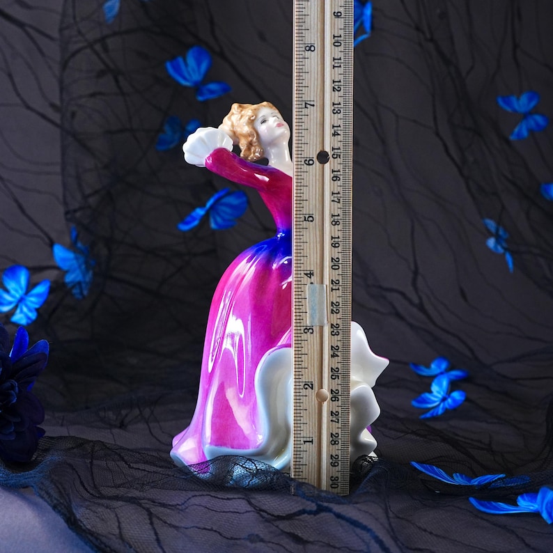 May include: A porcelain figurine of a person in a vibrant magenta and blue gown with a white ruffled hem, holding a white fan. A wooden ruler is placed vertically against the figurine, showing a height of approximately 29 centimetres.