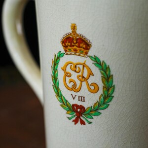 Puede incluir: Taza de cer&aacute;mica color crema con asa, con un emblema real. El emblema incluye una corona dorada, las letras "ER" en dorado, una corona de laurel verde y un lazo rojo. El n&uacute;mero romano "VIII" est&aacute; debajo.